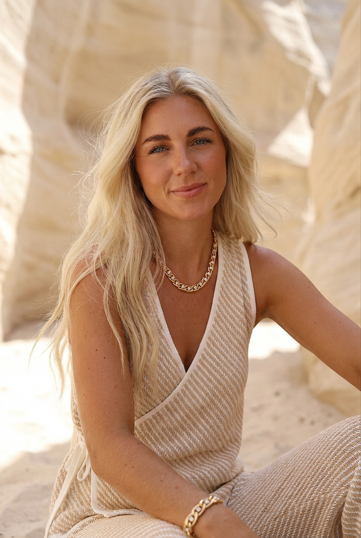 Woman in a beige dress sitting on sand with rock formations in the background with chunky gold necklace with diamond cz accents